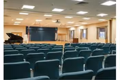 Interior view of a large room with rows of green upholstered chairs facing a stage area with a piano, podium, and a projection screen. The room has a drop ceiling with fluorescent lighting and windows along the right wall.