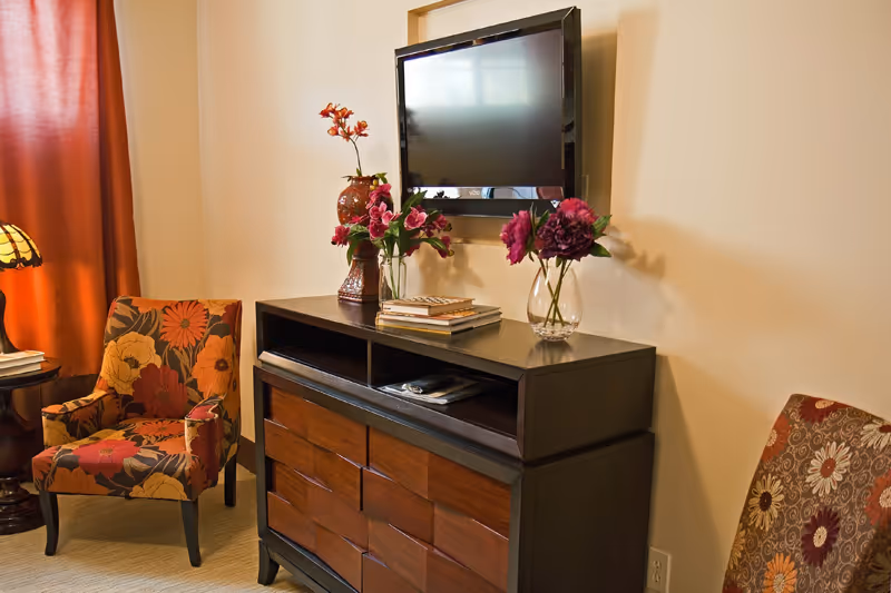 A cozy living room area with a dark wooden dresser topped with books and vases of flowers, a flat-screen TV mounted on the wall above it, and two upholstered armchairs with floral patterns, one in orange and yellow tones and the other in red and brown tones, next to a window with orange curtains.