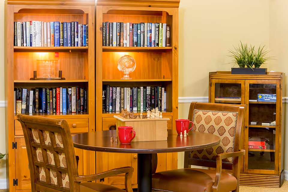A cozy reading and game area with two wooden bookshelves filled with books and decorative items, a round wooden table with a chess set and two red mugs, and two cushioned wooden chairs with patterned upholstery. A small cabinet with glass doors and a potted plant on top is visible in the background.