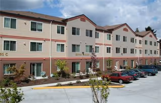Three-story beige and brick senior living building with landscaping and parked cars in front under a partly cloudy sky.