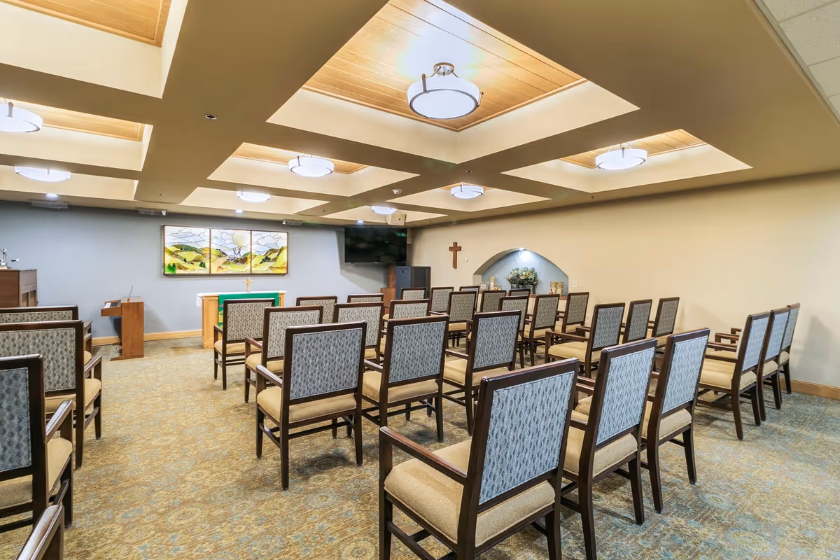A chapel room with rows of wooden chairs with patterned upholstery facing an altar. The altar has a green cloth and a cross, with a large stained glass window depicting a landscape behind it. The ceiling features recessed wooden panels with modern light fixtures. A cross is mounted on the wall to the right, and there is a small alcove with flowers and candles.
