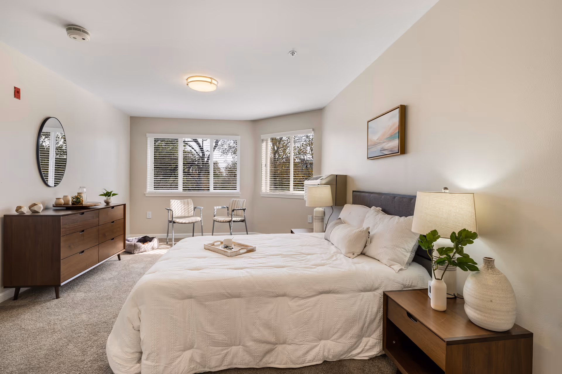 A bright and spacious bedroom with a large bed covered in white bedding. There is a wooden nightstand with a lamp and decorative vases on the right side of the bed. On the left side of the room, there is a wooden dresser with a round mirror above it and some decorative items. Two chairs are positioned near the windows, which have white blinds and offer a view of trees outside. The walls are painted light beige, and the room has carpeted flooring.