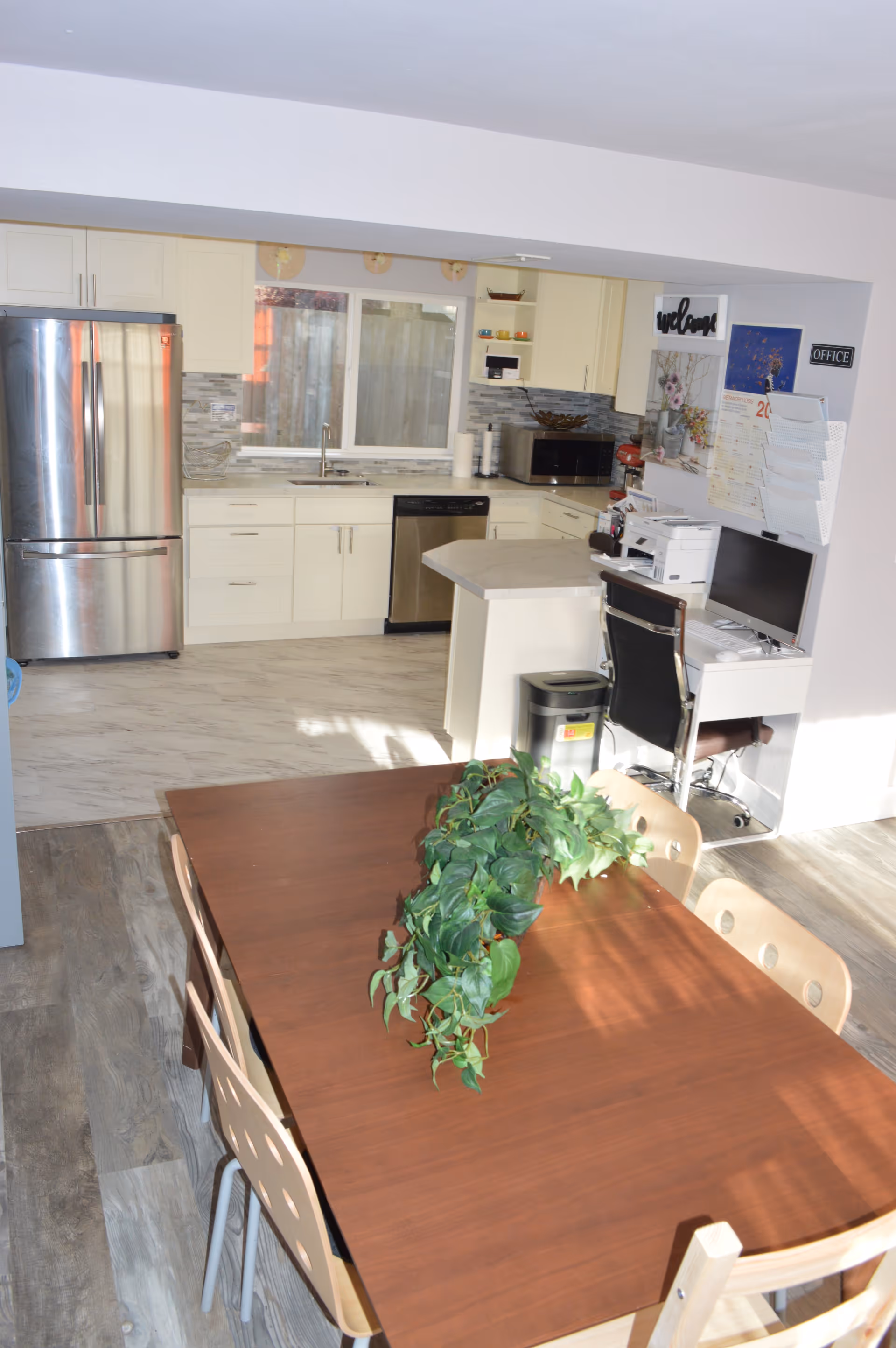 A bright kitchen and dining area featuring a wooden dining table with a green leafy plant centerpiece and four chairs. The kitchen has white cabinets, a stainless steel refrigerator, dishwasher, microwave, and a countertop with a small office workspace including a computer and printer. The floor is a mix of wood and tile, and there is a window above the sink.
