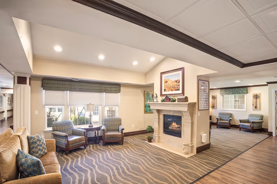 A cozy living room area in a senior living facility with a beige sofa, two patterned armchairs, a small wooden table with a lamp, and a fireplace with a framed picture above it. The room has soft lighting, beige walls, and a carpet with a wavy pattern. Another seating area with two armchairs is visible in the background.