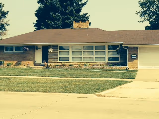 Single-story house with a brown roof, large front windows, a chimney, and a garage on the right side. The house is surrounded by a well-maintained lawn and a concrete driveway.