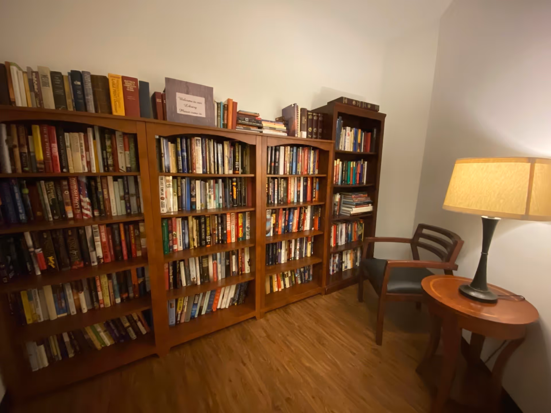 A cozy reading nook in a senior living facility featuring three wooden bookshelves filled with books, a wooden chair with armrests and a cushioned seat, and a round wooden side table with a lit table lamp. The room has wooden flooring and plain light-colored walls.