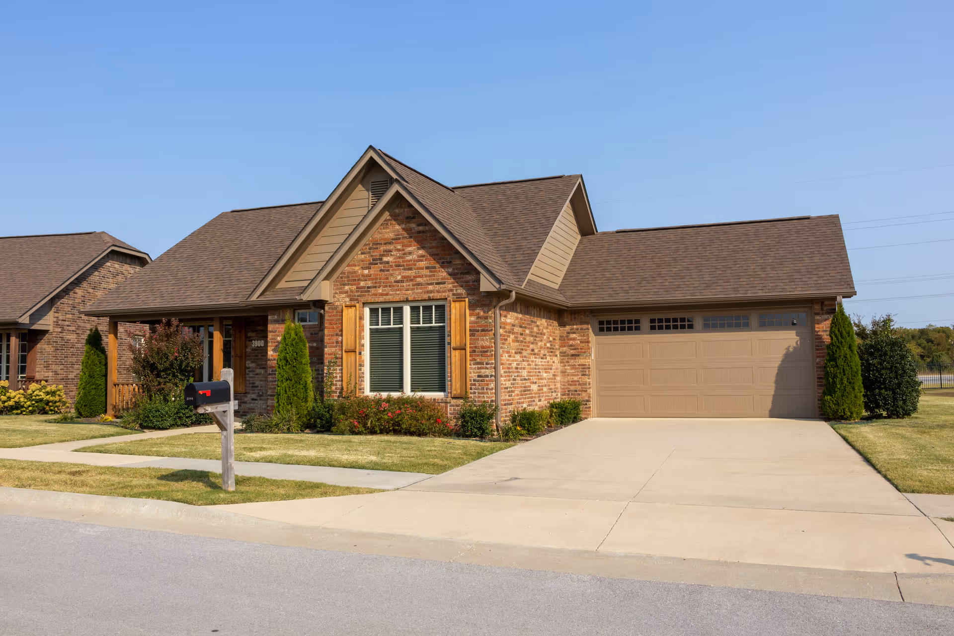 Single-story brick house with a two-car garage, driveway, mailbox and front lawn under a clear blue sky.
