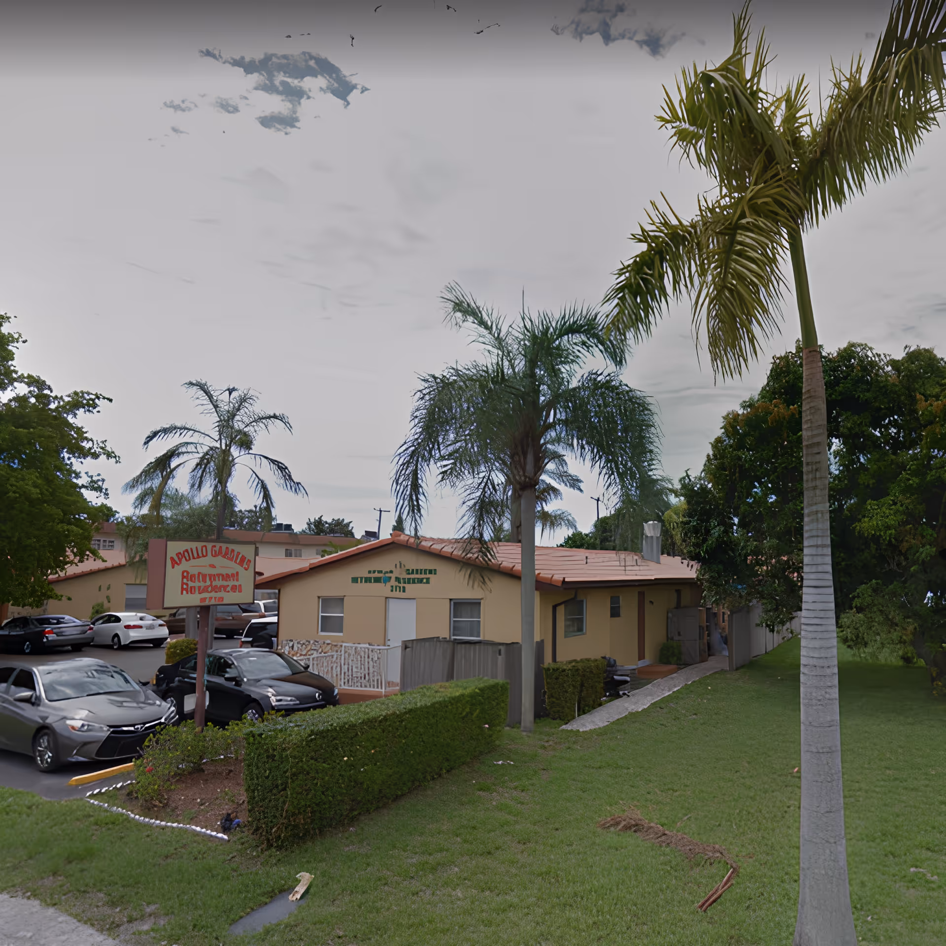 Exterior view of Apollo Gardens Retirement facility showing a single-story building with a red-tiled roof, surrounded by palm trees and greenery. Several cars are parked in the parking lot near the entrance, and a sign with the facility name is visible near the driveway.