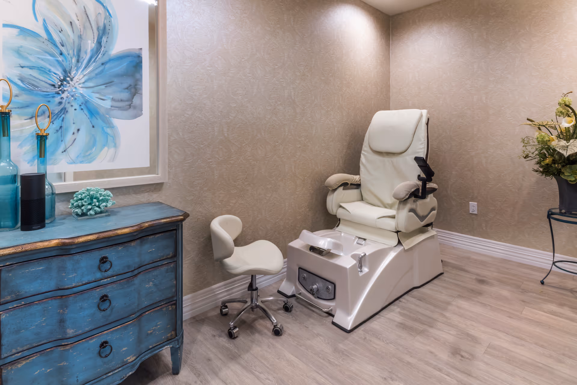 A pedicure spa chair with a foot bath basin and a small rolling stool in a softly lit room with beige patterned wallpaper and light wood flooring. To the left is a blue distressed wooden dresser with decorative items including blue glass bottles and a coral ornament, and a large framed artwork of a blue flower. To the right is a small table with a floral arrangement.