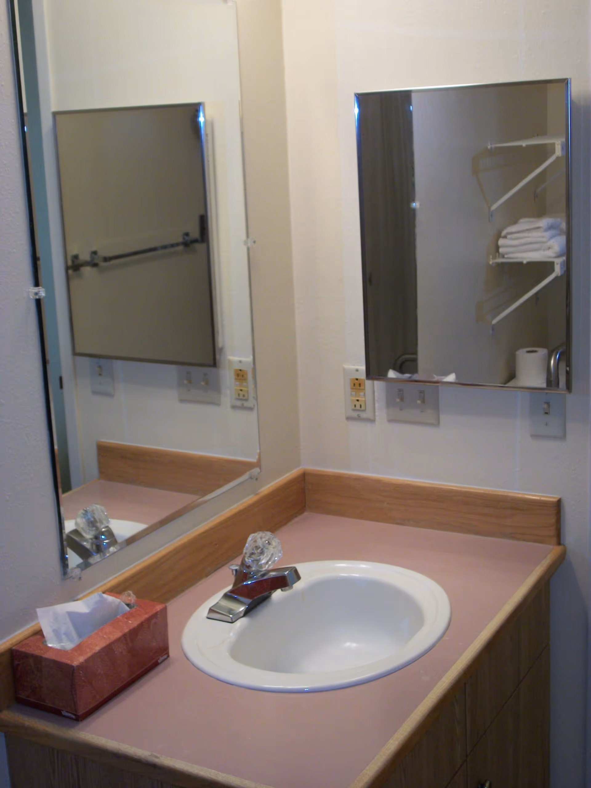 A bathroom sink with a pink countertop and wooden trim. There is a chrome faucet and a red tissue box on the counter. Two mirrors are mounted on the walls above the sink, one large rectangular mirror and one smaller mirrored medicine cabinet. In the reflection, shelves with folded white towels and a roll of toilet paper are visible.