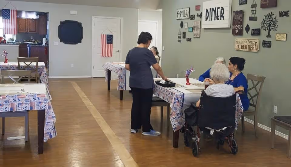 A dining area in an assisted living facility with tables covered in patriotic-themed tablecloths. Several elderly individuals are seated at one table, including one in a wheelchair, while a caregiver stands nearby attending to them. The walls are decorated with various signs and plaques, including one that says 'DINER' and another that says 'Friends & Family Gather Here'. An American flag hangs on a door in the background, and a kitchen area is visible through an open doorway.