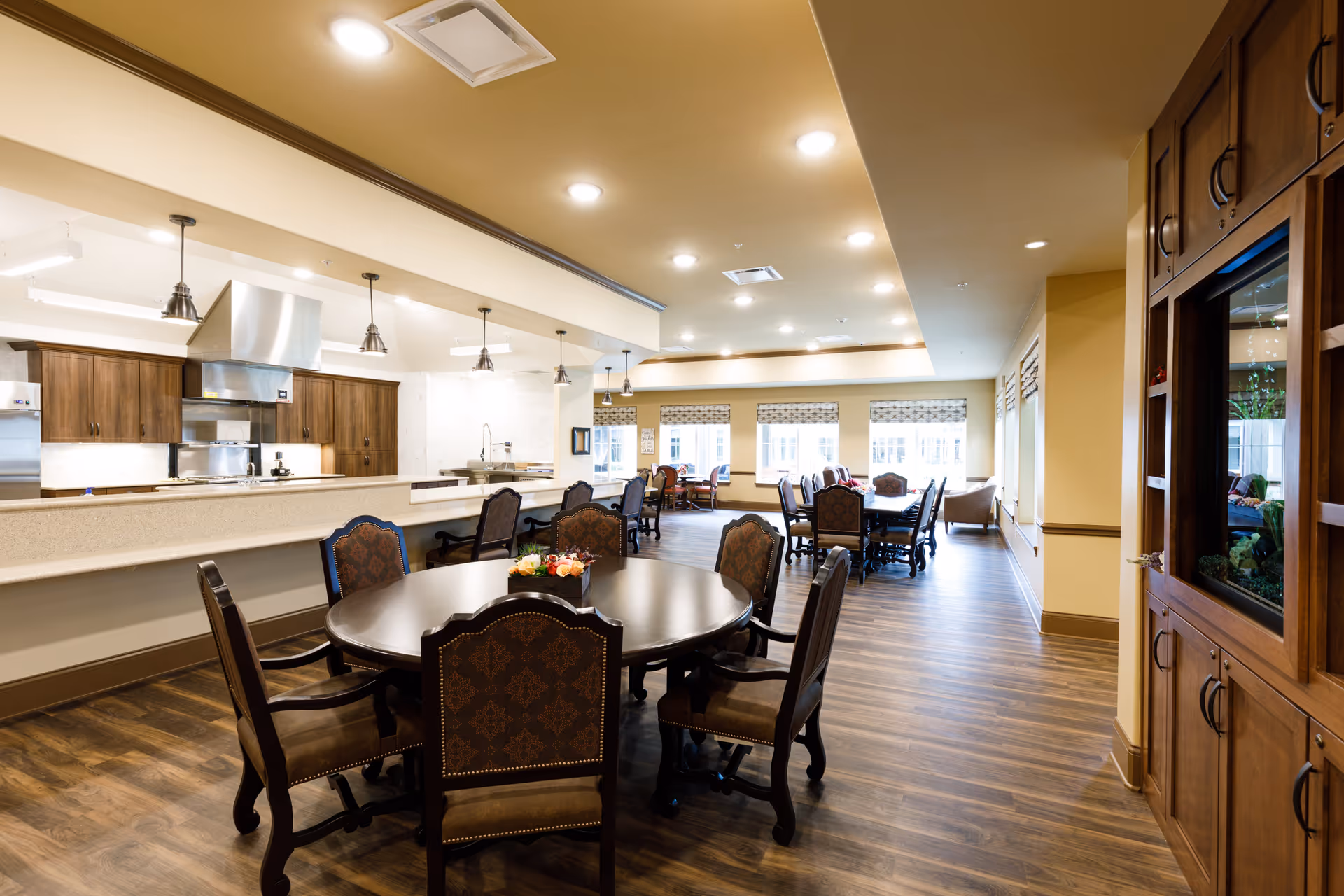 A spacious dining area in a senior living facility featuring multiple round and rectangular tables with cushioned chairs. The room has wooden flooring, warm yellow walls, and large windows with patterned blinds allowing natural light. The kitchen area with wooden cabinets, stainless steel appliances, and pendant lighting is visible in the background.