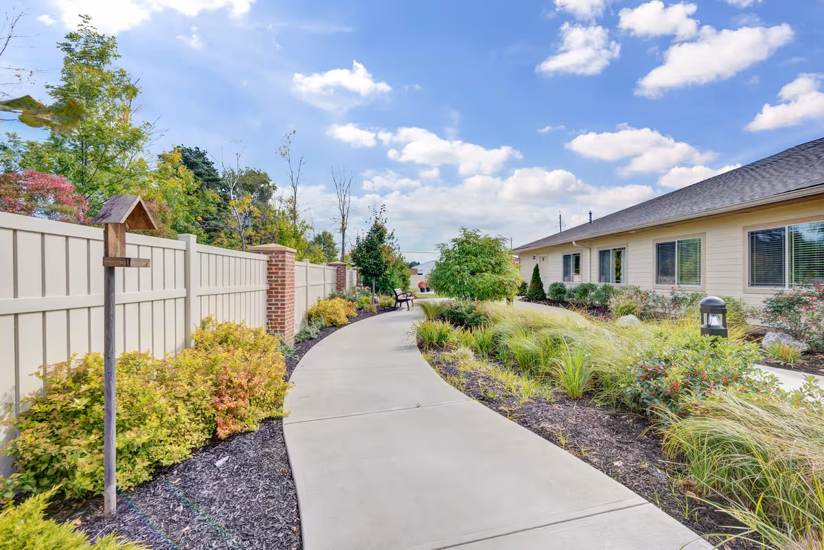 A curved concrete pathway runs through a landscaped garden area with various shrubs, grasses, and small trees. On the left side, there is a white fence with brick pillars and a wooden birdhouse mounted on a post. On the right side, there is a single-story building with beige siding and multiple windows. The sky is blue with scattered clouds.