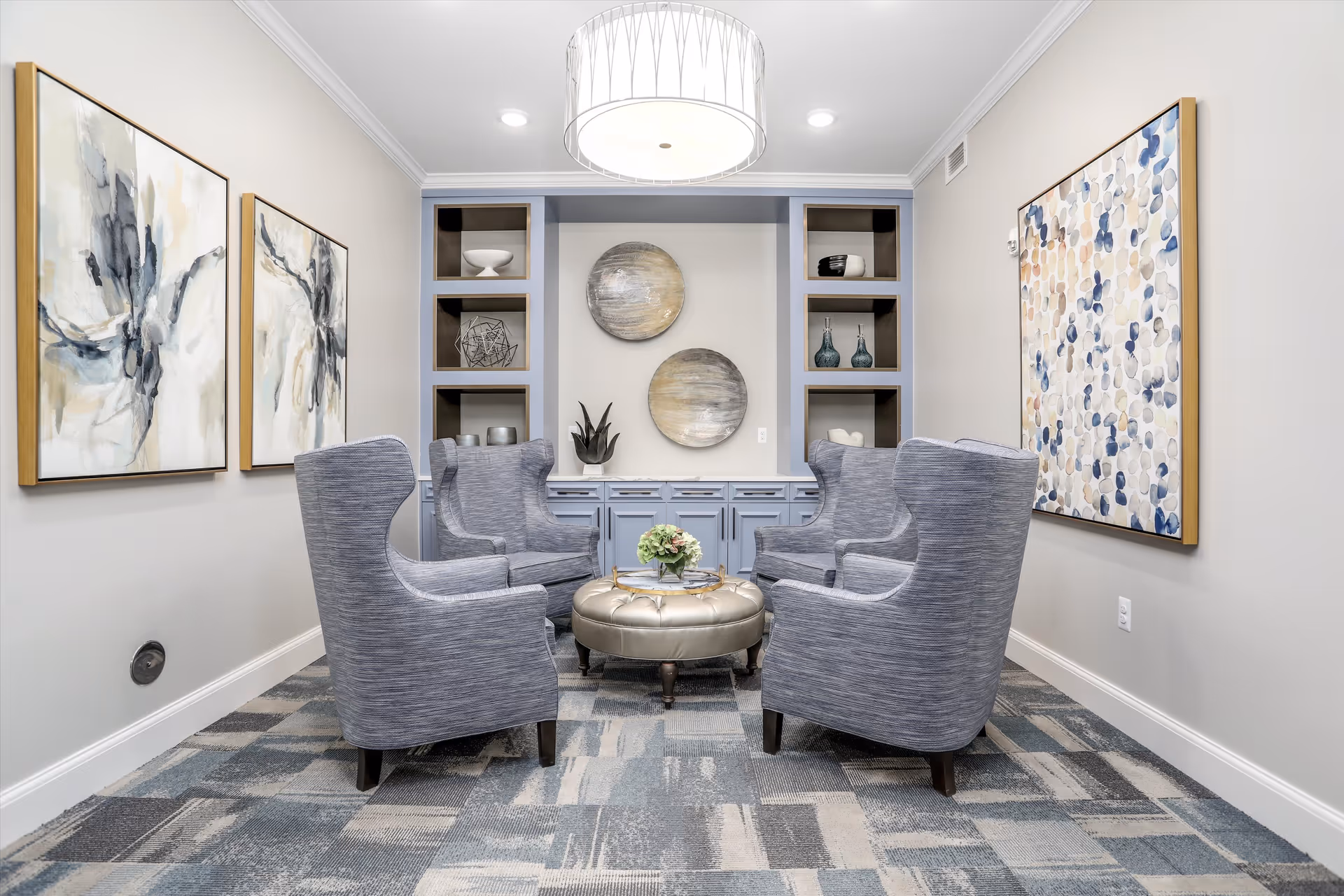 Small communal lounge with four gray wingback chairs arranged around a round ottoman, built-in shelving, and abstract wall art.