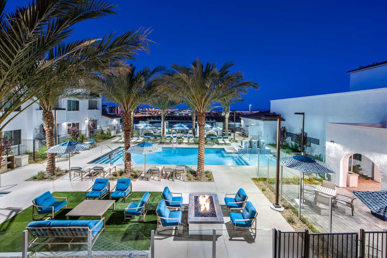 Outdoor pool area at night with palm trees, lounge chairs with blue cushions, umbrellas, and a fire pit surrounded by seating. White buildings and city lights are visible in the background.