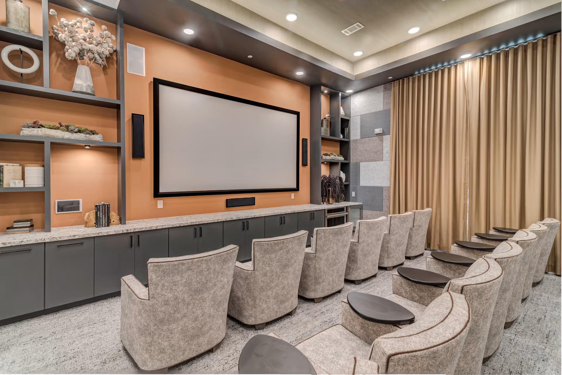 A modern home theater room with two rows of comfortable beige armchairs facing a large blank projection screen mounted on a warm orange wall. The room features built-in shelving with decorative items, recessed lighting in the ceiling, and beige curtains covering the windows on the right side.