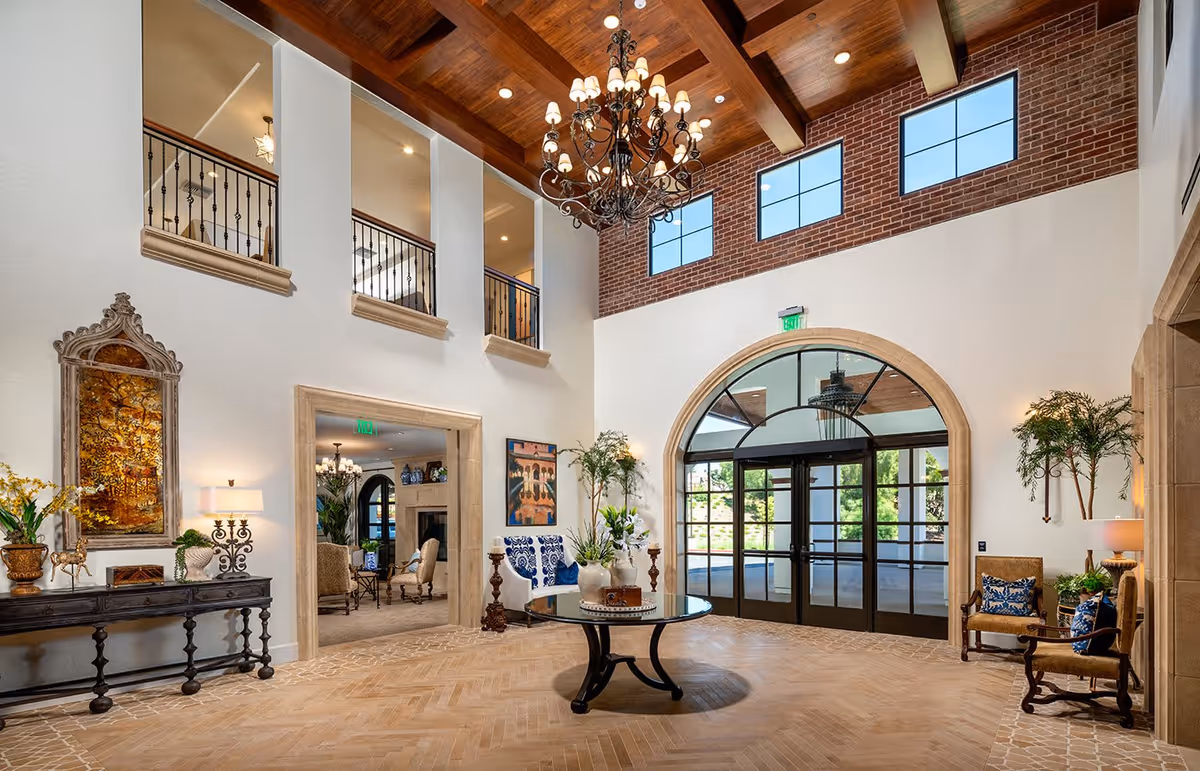 Spacious and elegant lobby area with high wooden ceiling, large chandelier, arched glass double doors, decorative plants, and comfortable seating. The walls feature brick accents and large windows allowing natural light to fill the space.