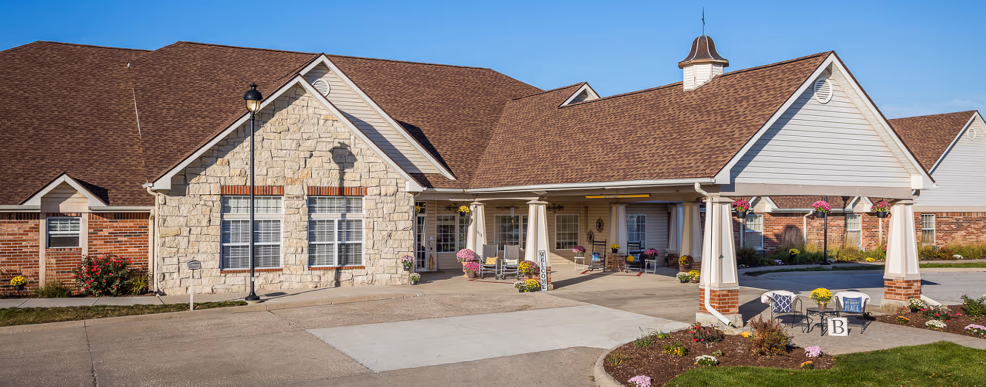 Exterior view of a single-story senior living facility building with a covered entrance supported by white columns. The building features a combination of stone and brick facade with a brown shingled roof. There are flower pots and landscaping around the entrance area under a clear blue sky.