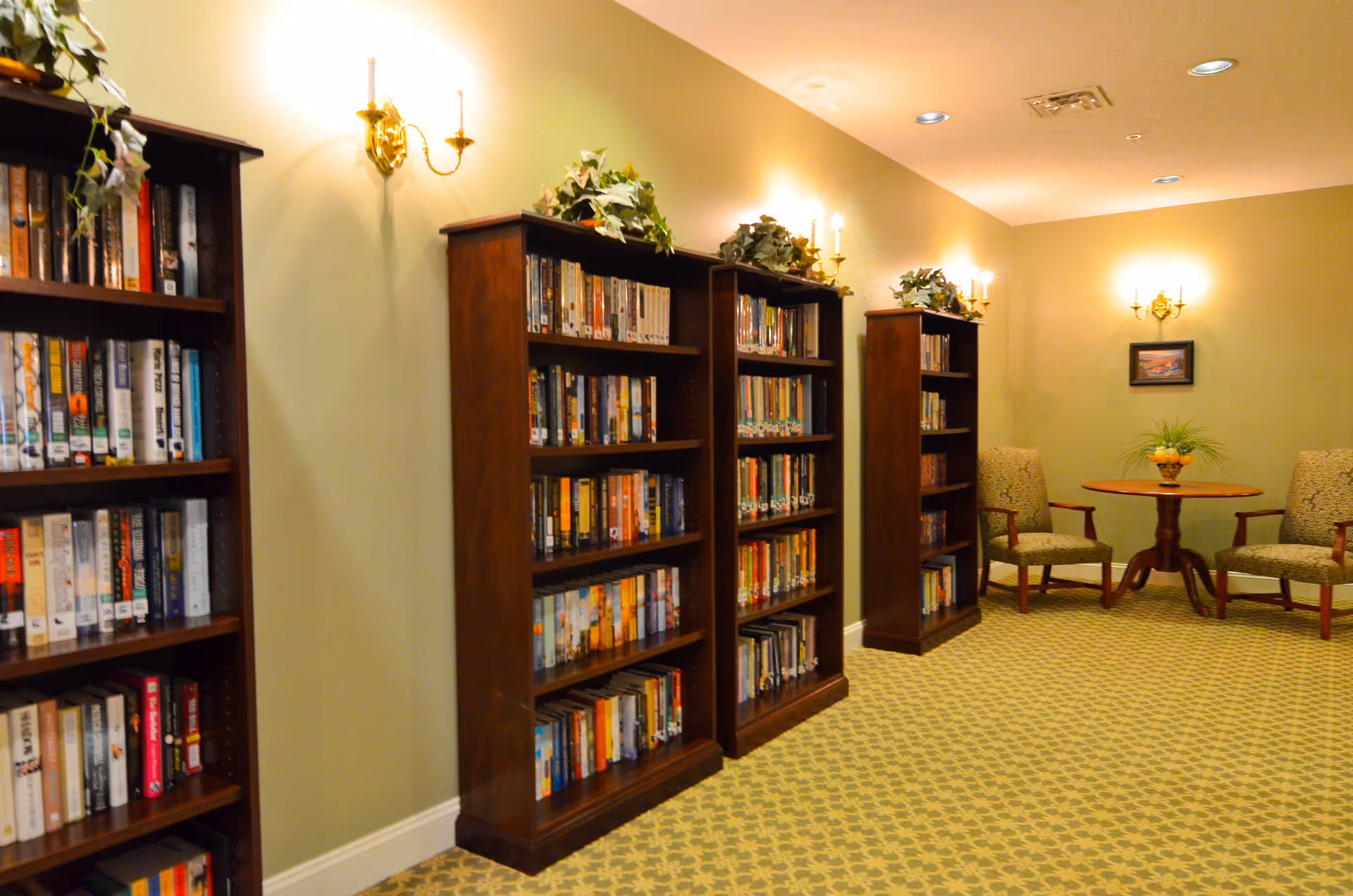 Reading area with wooden bookshelves filled with books, wall sconces, patterned carpet, and a small table flanked by two chairs.