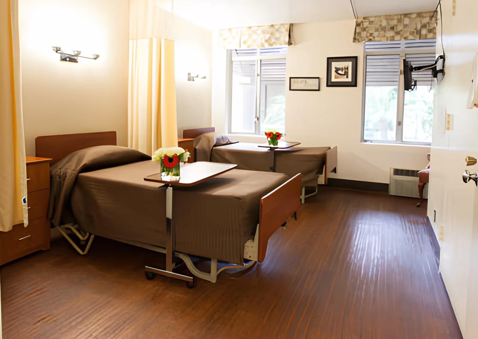 A room with two hospital-style beds covered with brown bedspreads, each with a small overbed table holding a vase of flowers. The room has wooden flooring, two windows with patterned valances, framed pictures on the wall, and a wall-mounted TV. There are bedside tables next to each bed and a curtain partially drawn between the beds.