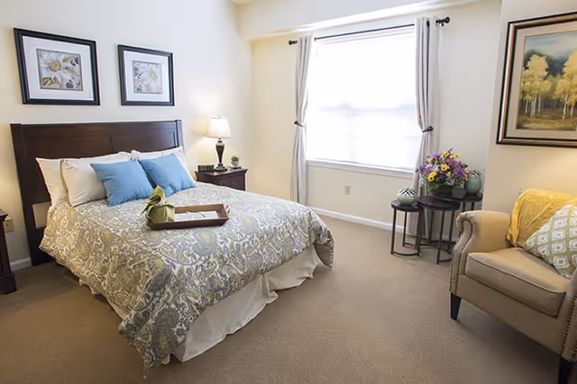 Bright furnished bedroom with a made bed, nightstand and lamp, armchair, and a window with curtains.