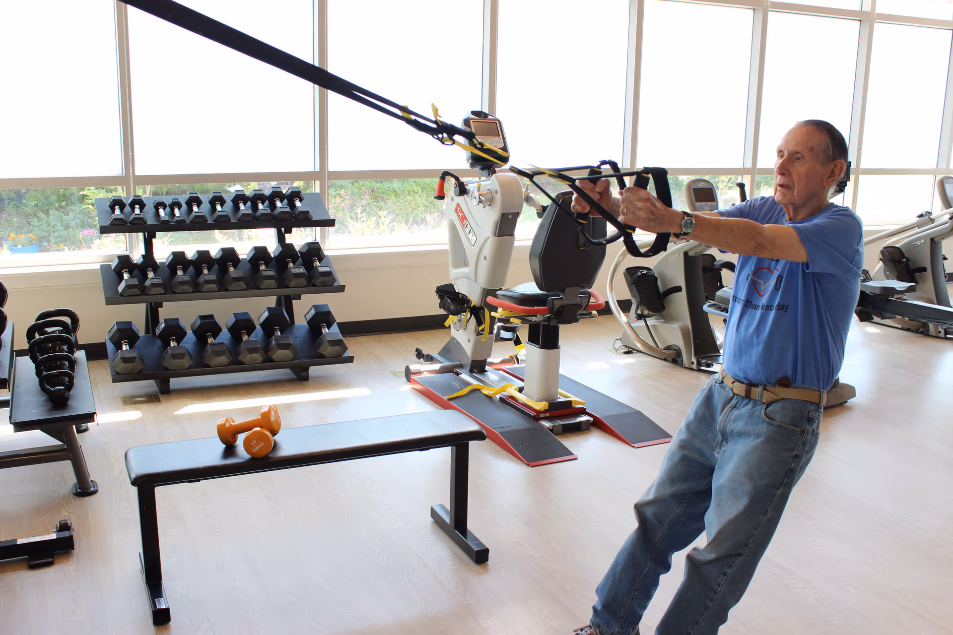 An elderly man exercising in a fitness room using resistance bands attached to a stationary machine. The room has large windows letting in natural light, a rack of dumbbells, kettlebells, and other exercise equipment including stationary bikes and a bench with orange dumbbells on it.