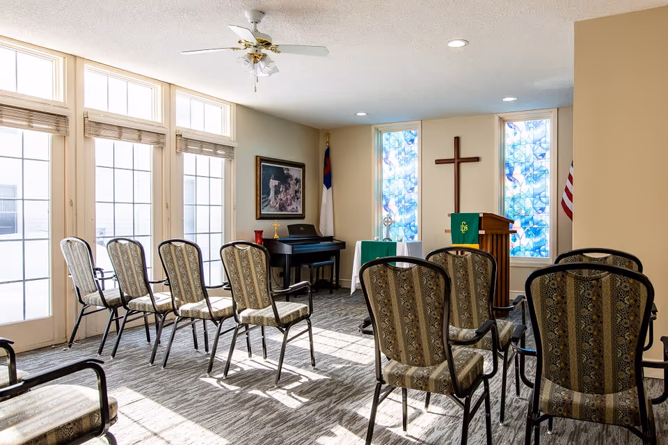 Sunlit chapel-style room with rows of patterned chairs facing a podium, wooden cross, stained glass windows and a piano.