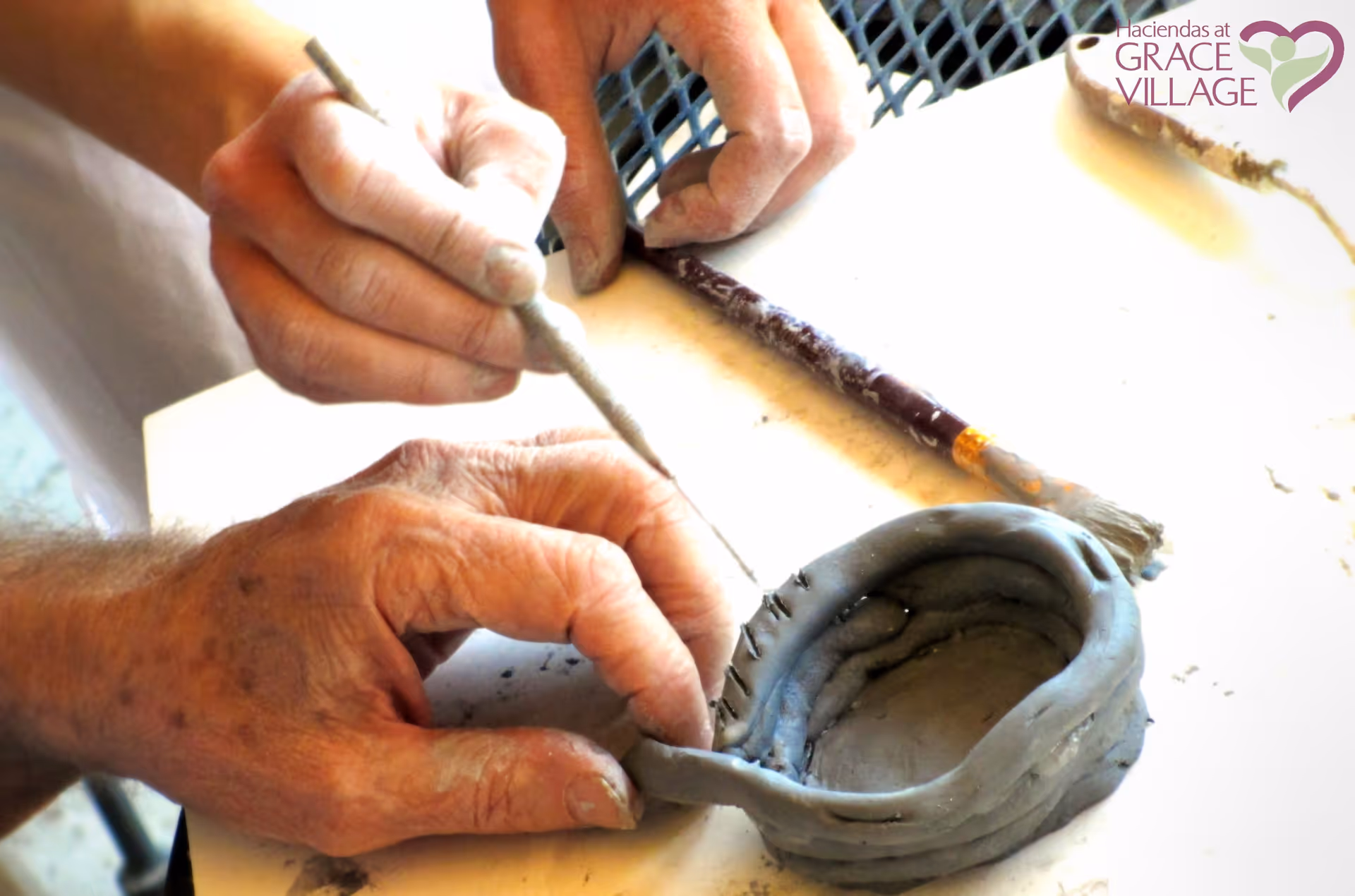 Close-up of two pairs of hands working on shaping and detailing a small clay pot on a table, with clay sculpting tools nearby.