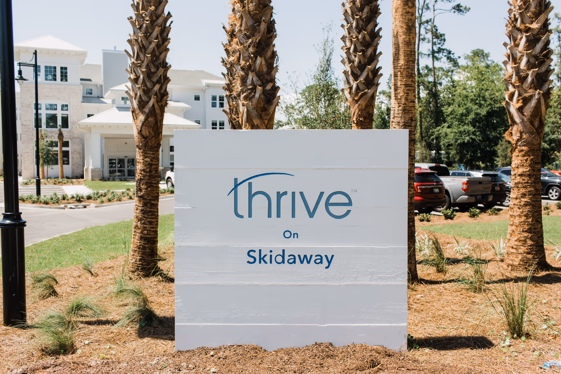 A white wooden sign with the text 'thrive On Skidaway' in blue letters, placed outdoors among palm trees and landscaping. In the background, there is a large white building and a parking lot with several cars.