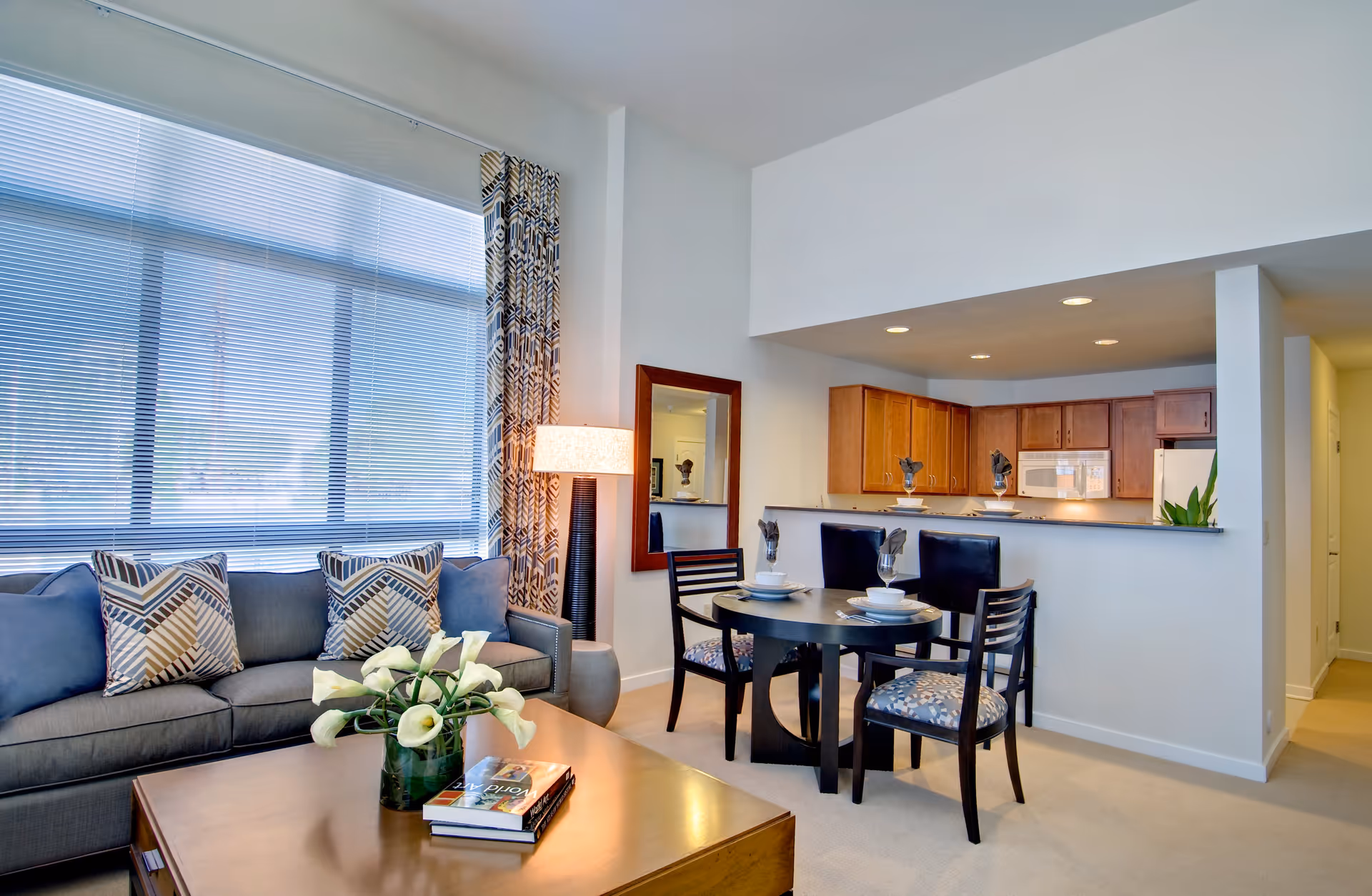 A bright and modern living room and dining area with a large window covered by blinds and patterned curtains. The living room has a gray sofa with patterned cushions, a wooden coffee table with a vase of white flowers and books. The dining area features a round table set with plates, bowls, and glasses, surrounded by four chairs. The kitchen is visible in the background with wooden cabinets, a microwave, and a refrigerator. A tall floor lamp and a wall mirror are also present.