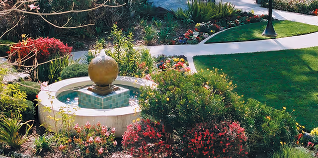 A small decorative fountain surrounded by colorful flower beds, shrubs, a lawn and winding walkways in a landscaped garden.