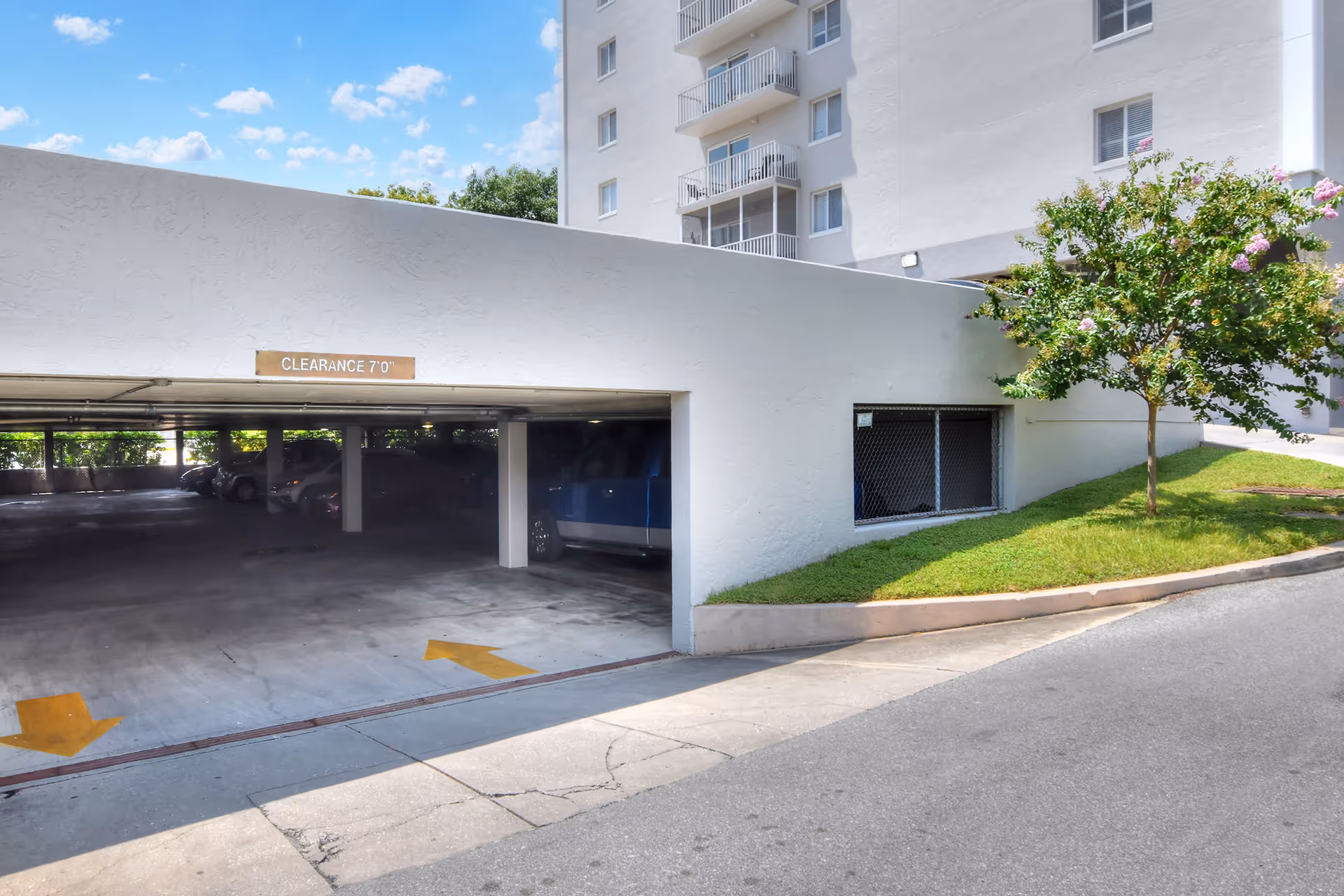 Covered parking garage entrance beneath a white apartment building with a clearance sign and a small tree on a grassy ramp.