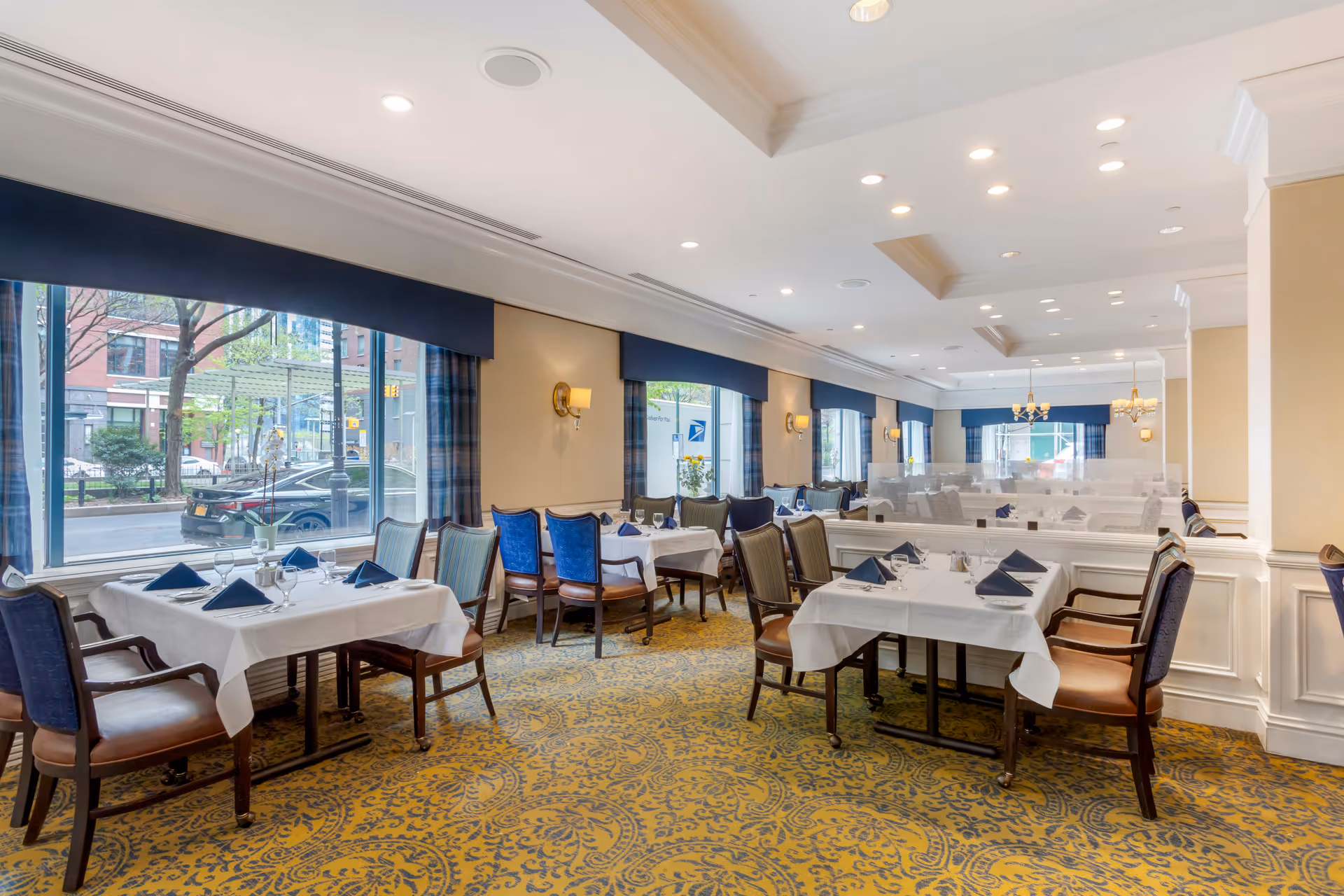 A bright and elegant dining room with multiple tables covered in white tablecloths, each set with blue napkins, glassware, and silverware. The room features large windows with blue valances, patterned carpet, and wall sconces providing warm lighting. Outside the windows, a street and buildings are visible.