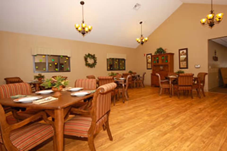A dining room in a senior living facility with wooden tables and striped upholstered chairs. The tables are set with plates and napkins, and there are potted plants as centerpieces. The room has wooden flooring, beige walls, two windows with valances, a wreath decoration, framed pictures, and a wooden cabinet. Three chandeliers with yellow lights hang from the ceiling.
