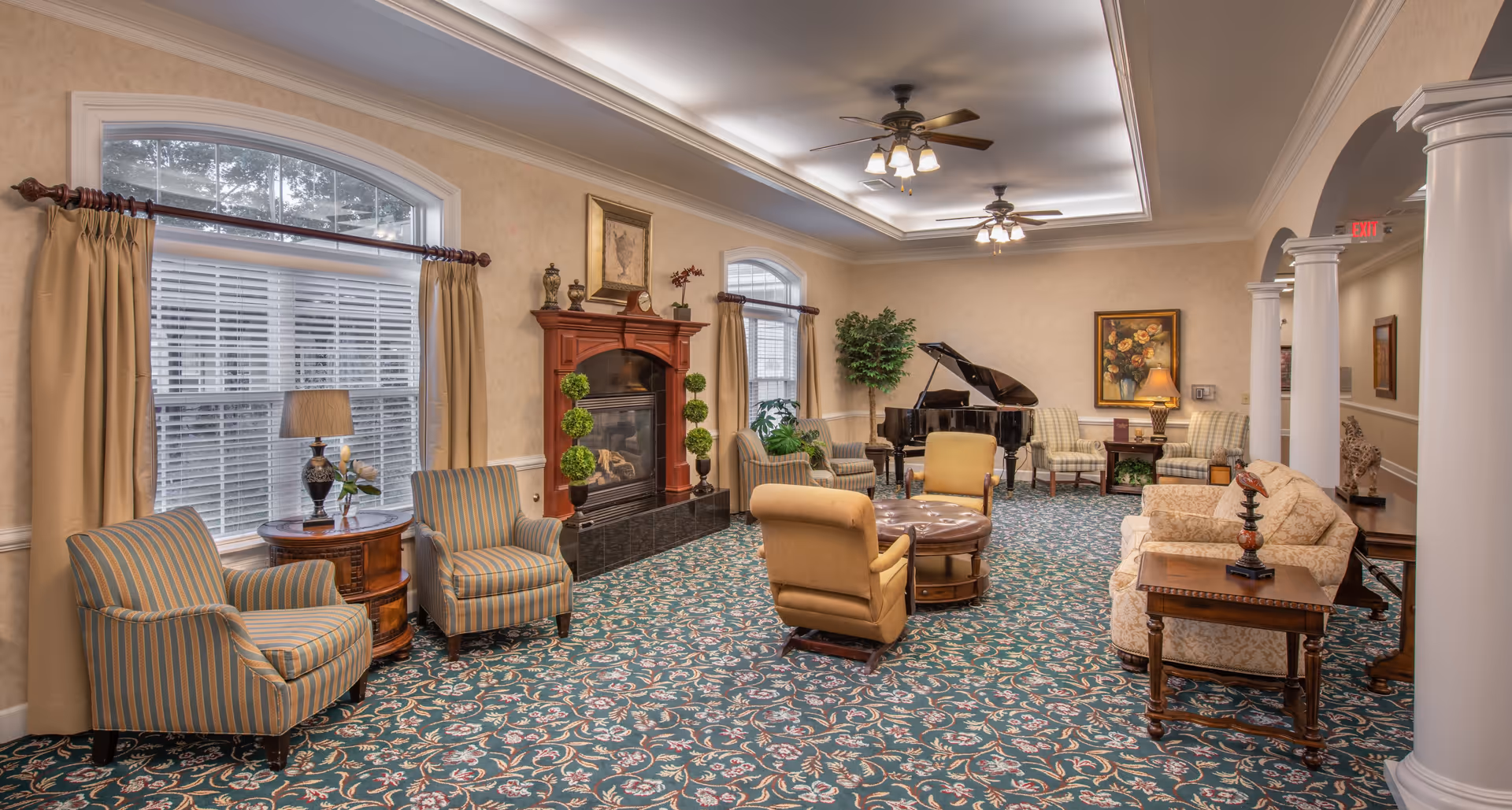 A spacious and elegantly decorated living room with patterned carpet, multiple upholstered chairs and sofas arranged around a central coffee table, a fireplace with decorative plants on either side, large windows with curtains, a grand piano in the corner, ceiling fans with lights, and framed artwork on the walls.