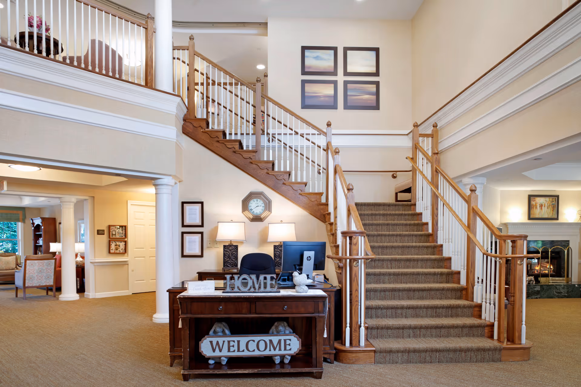 Interior view of a senior living facility lobby with a carpeted staircase featuring wooden handrails and white balusters. At the base of the stairs is a wooden desk with a computer, two lamps, and decorative signs that say 'HOME' and 'WELCOME'. The walls are beige with white trim, and there are framed pictures and a clock on the wall. To the left, there is a seating area with chairs and a window, and to the right, a fireplace with artwork above it.
