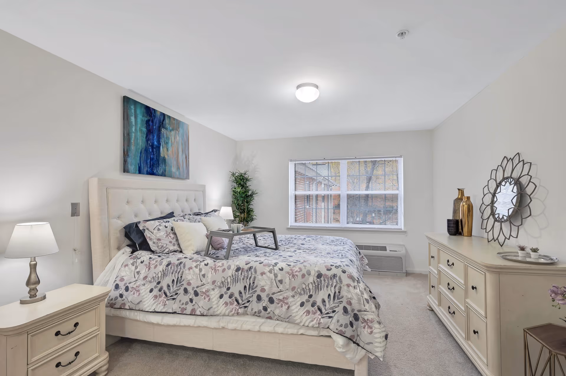 A bright and cozy bedroom with a large bed featuring a floral patterned comforter and multiple pillows. There is a small tray on the bed, a nightstand with a lamp on the left, and a dresser with decorative vases and a round mirror on the right. A large window with blinds lets in natural light, and a potted plant is placed in the corner near the window.