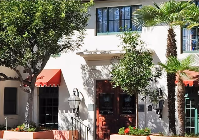 Exterior view of a building entrance with double wooden doors, surrounded by trees and plants including palm trees. The building has white walls, black framed windows, and red awnings above some windows.