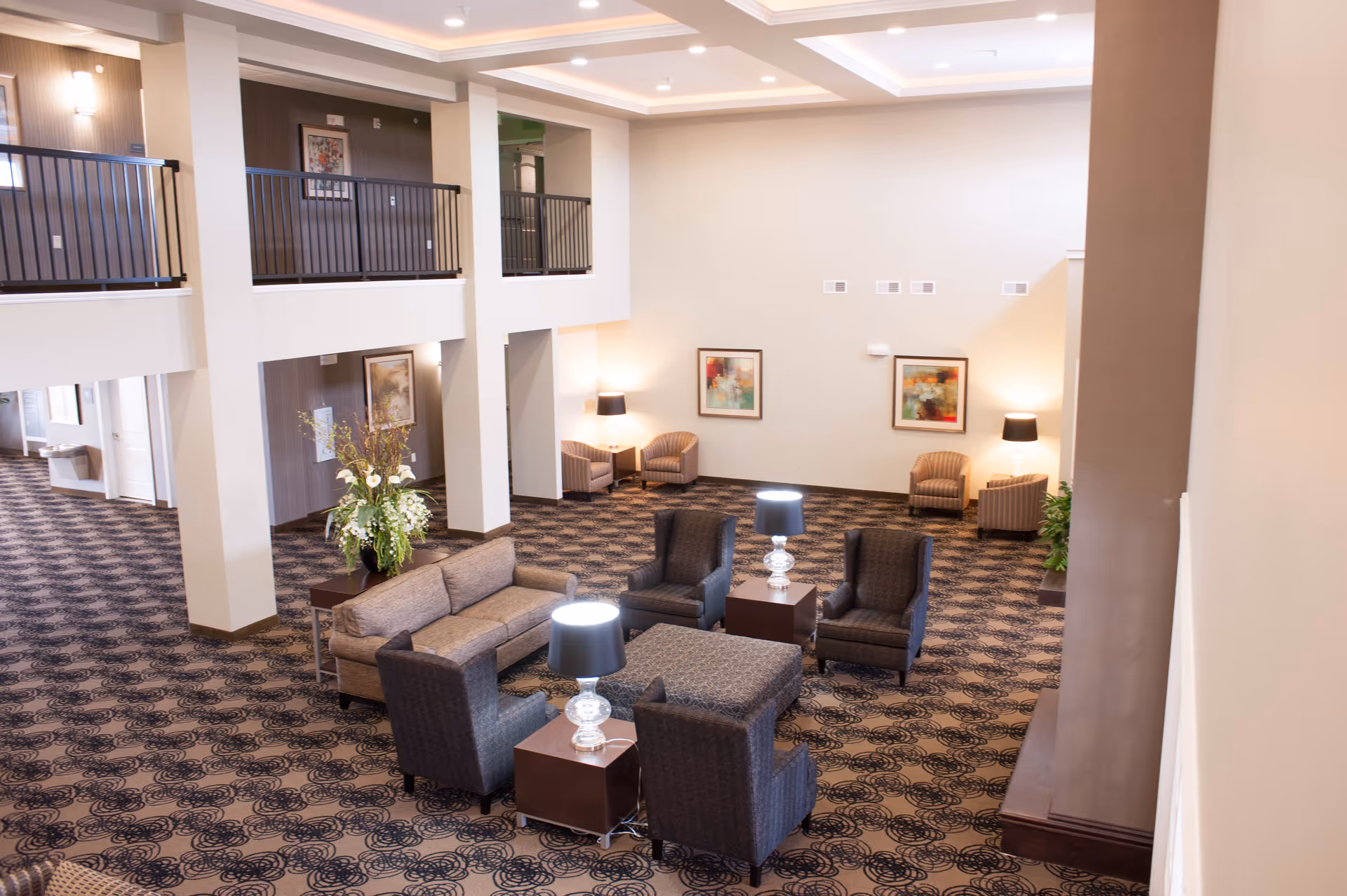 Spacious and well-lit common area with patterned carpet flooring, multiple seating arrangements including sofas and armchairs, side tables with lamps, decorative plants, and framed artwork on the walls. The area has a high ceiling with recessed lighting and a second-floor balcony overlooking the space.