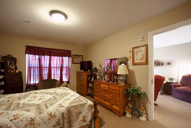 A cozy bedroom with a bed covered in a floral quilt, a wooden dresser with a lamp and decorative items, a window with red curtains, and an armchair near the window. The room has beige walls and carpeted floor. An open door leads to a living room with purple upholstered chairs and a lamp.