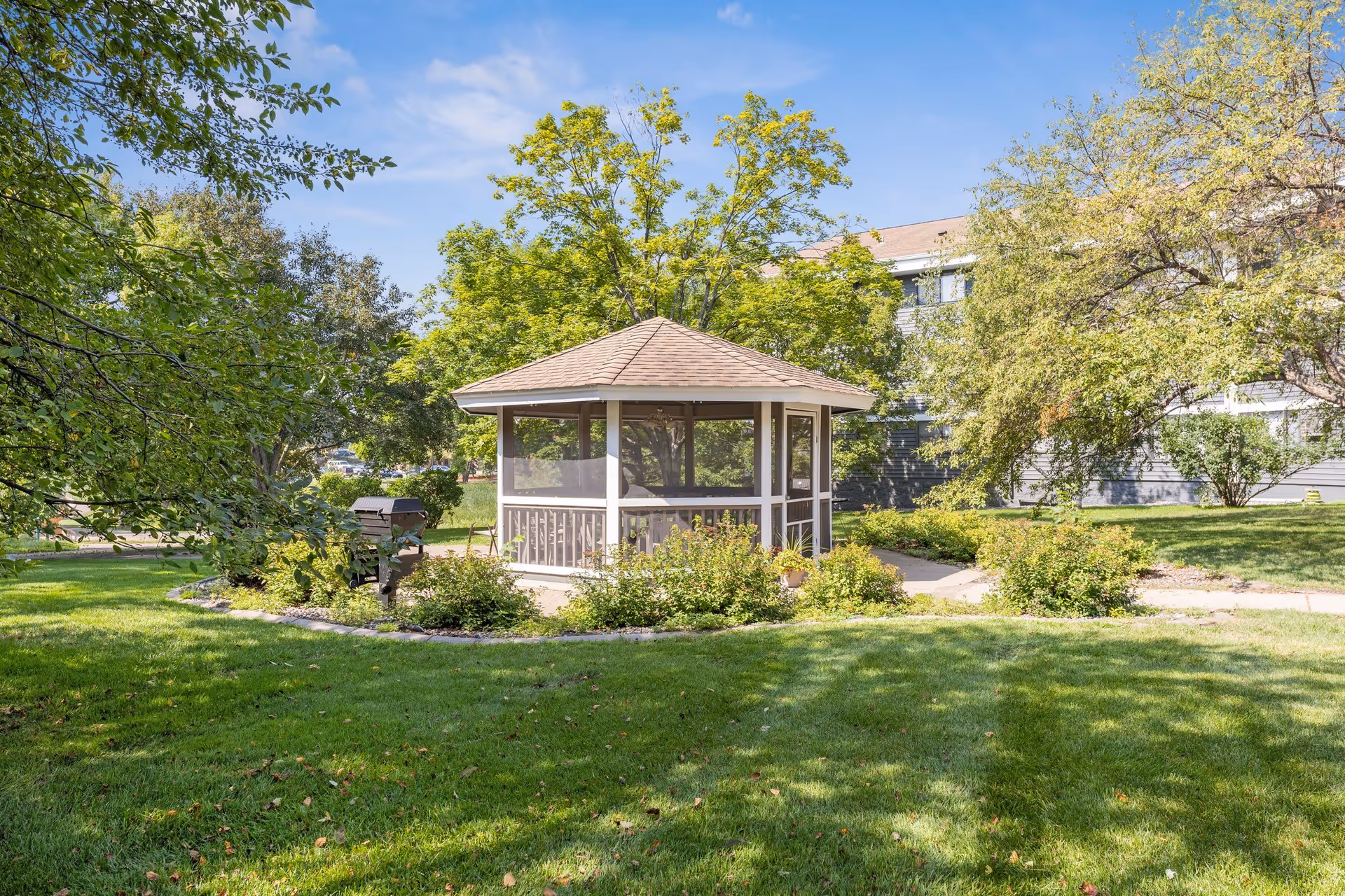 A small screened gazebo surrounded by green bushes and trees in a grassy outdoor area with a building partially visible in the background under a clear blue sky.