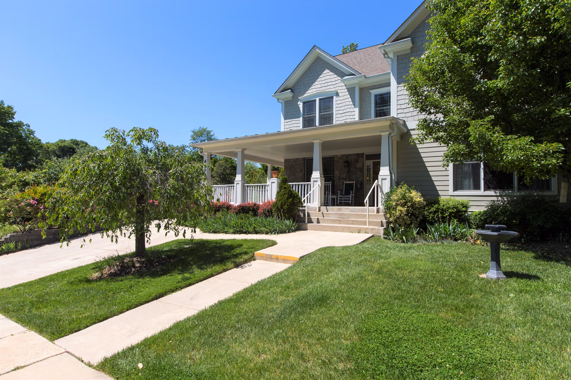 Front exterior view of a two-story house with a covered porch, white railings, and steps leading up to the entrance. The house is surrounded by a well-maintained lawn, trees, shrubs, and a birdbath on the right side. The sky is clear and blue.