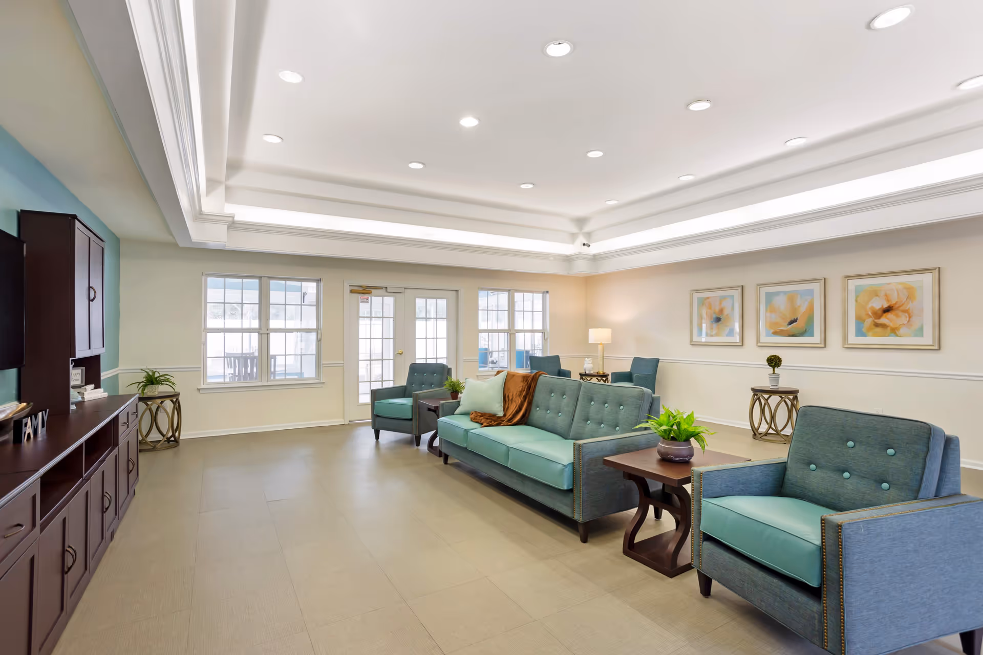 A bright and spacious living room area with teal upholstered sofa and armchairs, wooden side tables with potted plants, a dark wooden cabinet along the left wall, three framed floral paintings on the right wall, and large windows and glass doors letting in natural light.