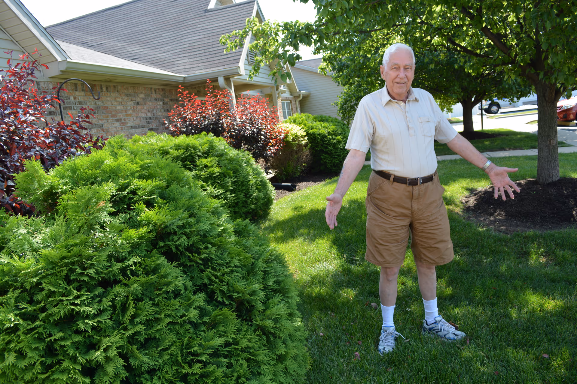 An elderly man standing outside on a green lawn with bushes and trees around him, in front of a brick building with a sloped roof. He is wearing a light-colored short-sleeve shirt, brown shorts, white socks, and sneakers, with his arms open wide and smiling.