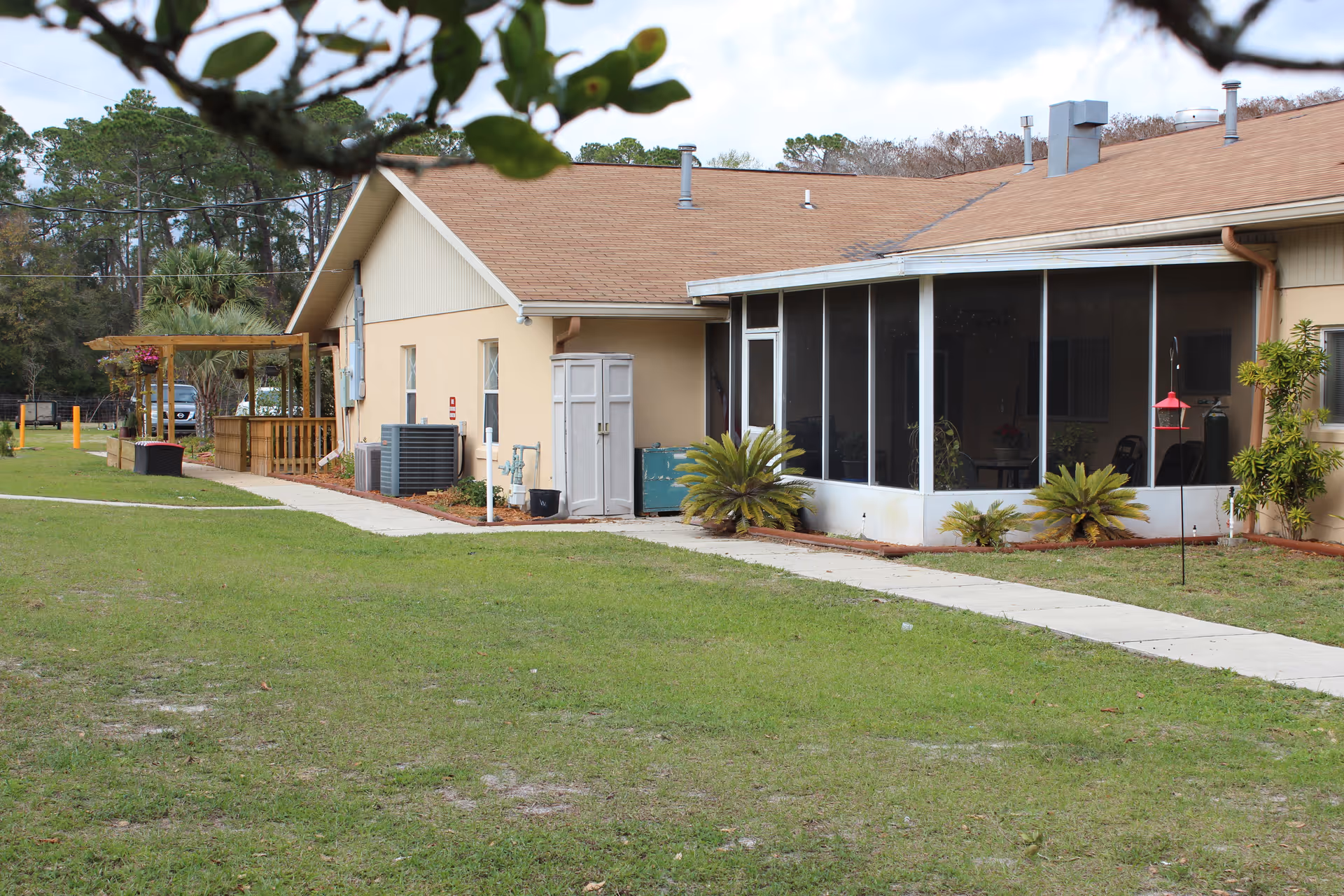 Exterior view of a single-story building with a brown roof and beige walls, featuring a screened porch area, a wooden pergola, and a concrete walkway surrounded by green grass and some small plants.