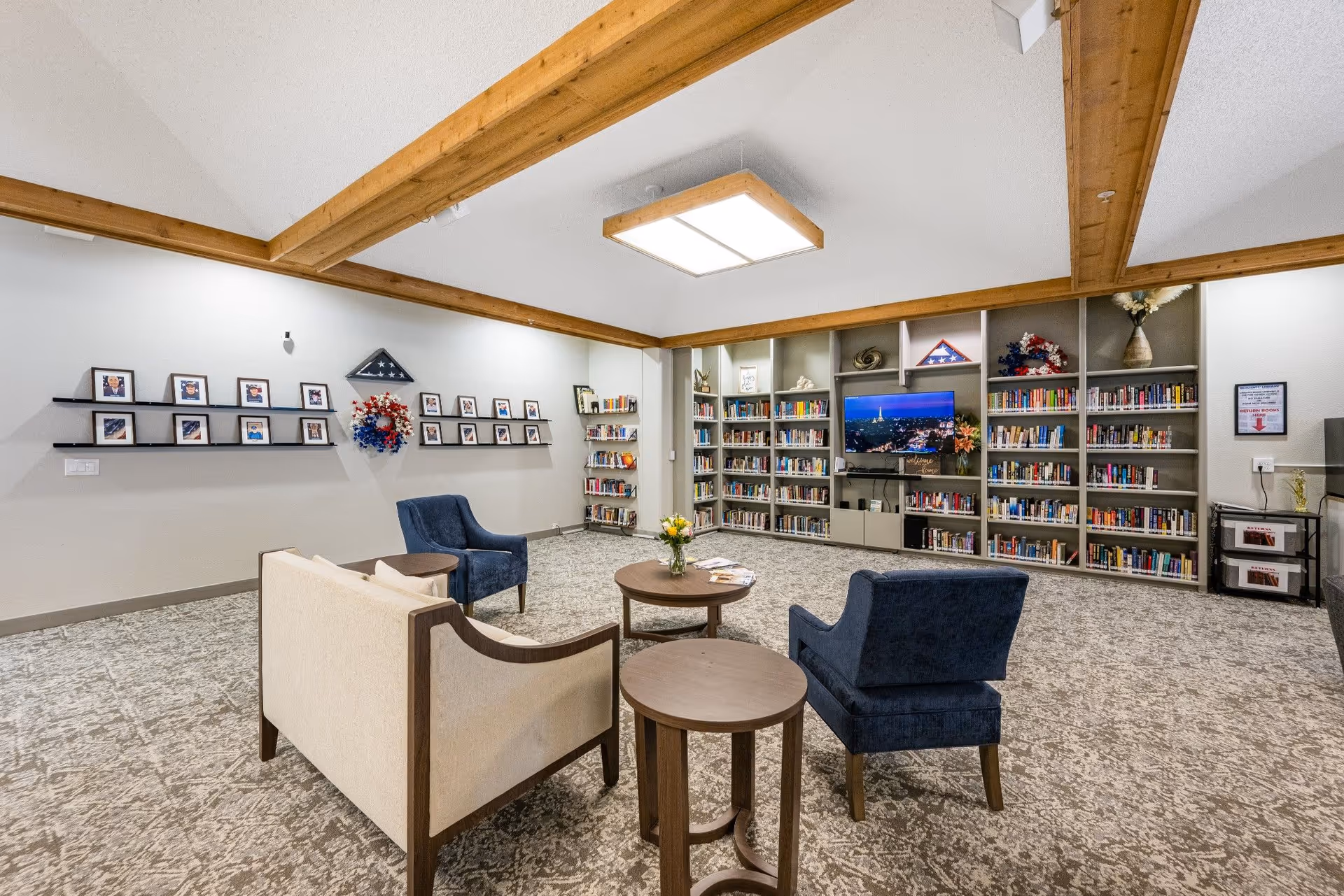 A cozy library room with beige carpet and wooden ceiling beams. There are bookshelves filled with books along the back wall, a flat-screen TV displaying a cityscape, and framed photos and patriotic decorations on the walls. The seating area includes a beige sofa, two blue armchairs, and two round wooden tables, one with a vase of flowers.