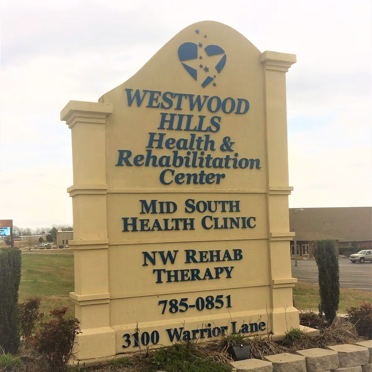 A large beige freestanding sign with blue lettering reading 'Westwood Hills Health & Rehabilitation Center' plus clinic, therapy, phone number, and address information in front of the facility.