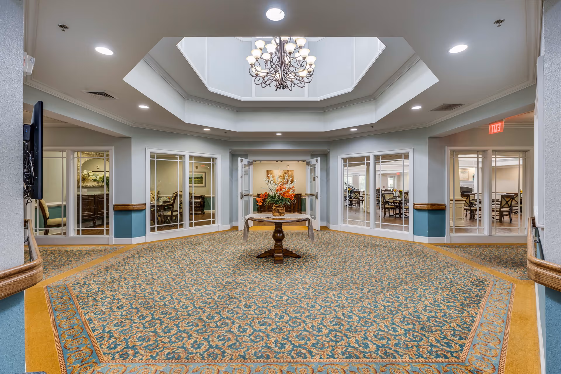Interior view of a senior living facility hallway with a patterned carpet and a round table with a floral arrangement in the center. The ceiling features a large skylight with a decorative chandelier. Glass doors and windows on the walls reveal dining areas with tables and chairs.