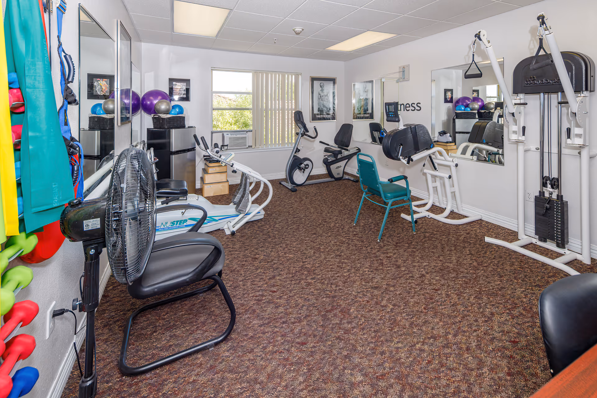 A small fitness room with exercise equipment including stationary bikes, a rowing machine, a weight machine, and various exercise balls and resistance bands. The room has carpeted flooring, white walls, large mirrors, and a window with vertical blinds letting in natural light.