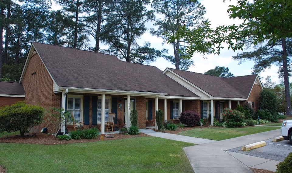 Exterior view of a single-story brick building with a brown shingled roof, white columns supporting a covered porch with rocking chairs, surrounded by green grass, shrubs, and tall pine trees under a partly cloudy sky.